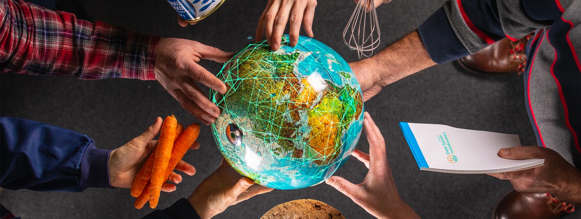 Keskellä maapallo, jonka ympärillä on käsiä. Osassa käsistä on tuotteita kuten porkkanoita, lehtiö, vispilä.