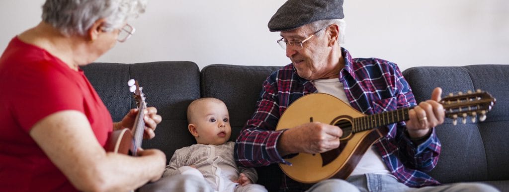 Ikääntynyt nainen ja mies istuvat sohvalla ja soittavat mandoliiniä. Heidän välissään istuu vauva kuunnellen.