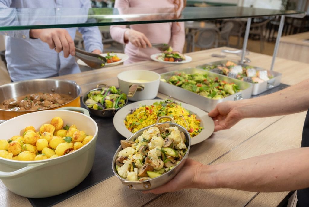 Kuvassa on buffet pöytä, jossa ruokia. Ympärillä ruokapalvelutyöntekijä sekä kaksi asiakasta.