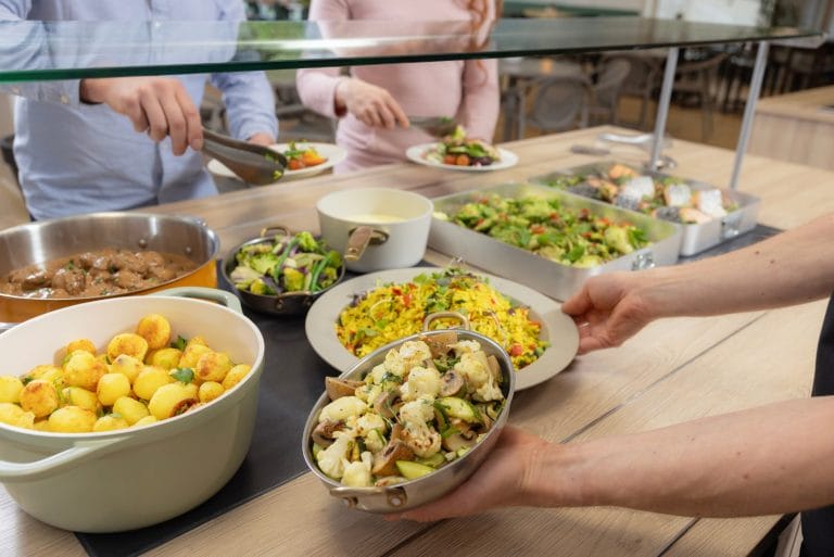Kuvassa on buffet pöytä, jossa ruokia. Ympärillä ruokapalvelutyöntekijä sekä kaksi asiakasta.