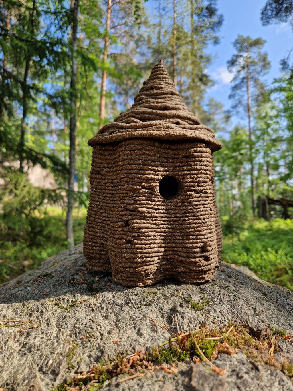 Biomateriaalista 3D-tulostettu linnunpönttö nököttää kivellä, taustalla puita.