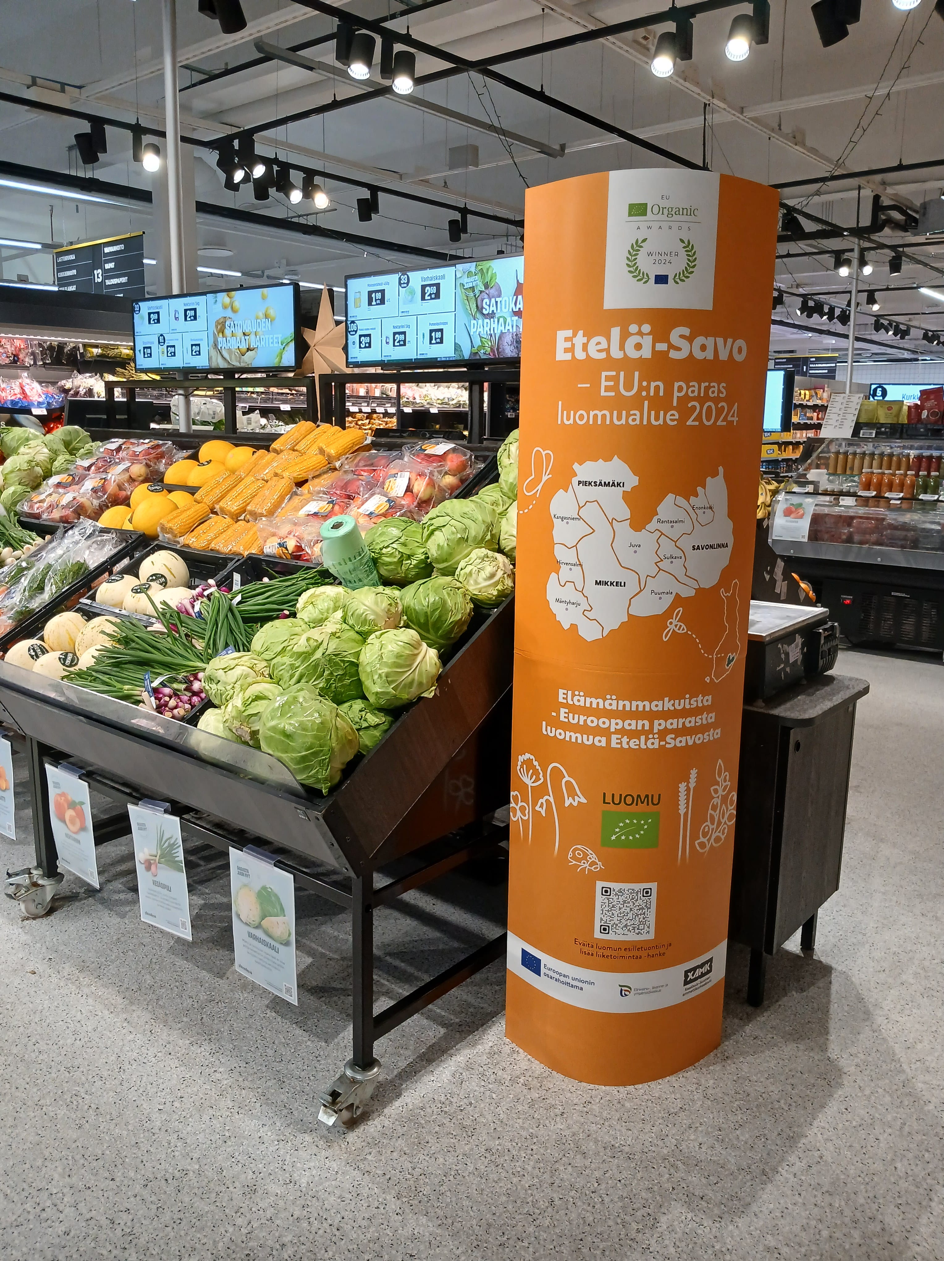 Kaupan vihannesosastolta kuva, jossa vihanneshyllyn päässä keltainen korkea mainos. Mainoksessa tekstiä ja logoja.