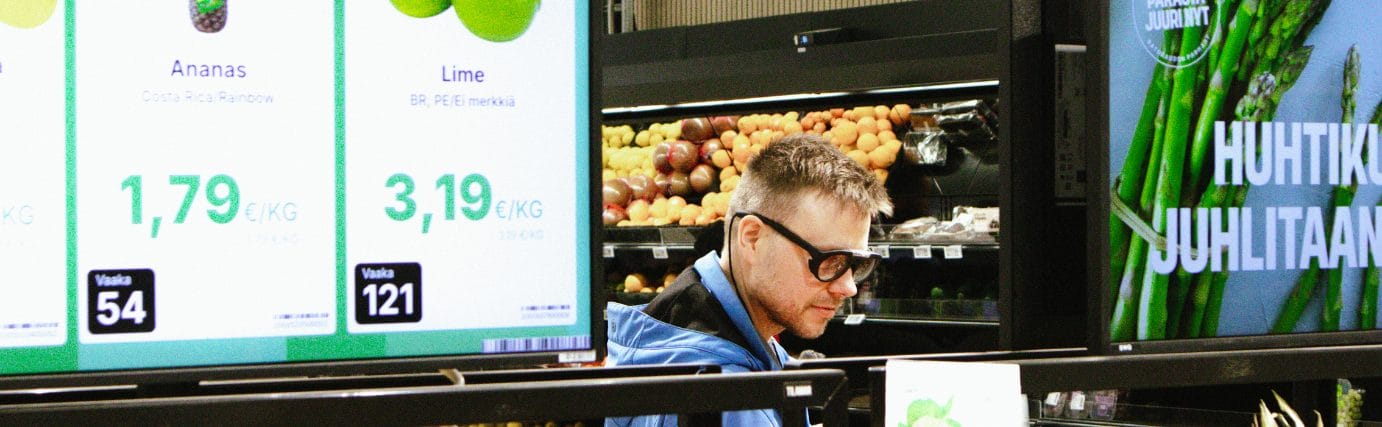 Mies on ruokakaupan hedelmä- ja vihannesosastolla puettava silmänliikekamera päässään. Etualalla näkyy digitaaliset hintanäytöt. Kuva: Heta Hotokka.