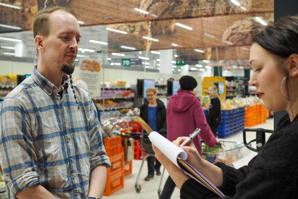 Nainen haastattelee miestä ruokakaupassa, taustalla näkyy myymälä ja muita asiakkaita ruokaostoksilla. Kuva: Sanni Siilin