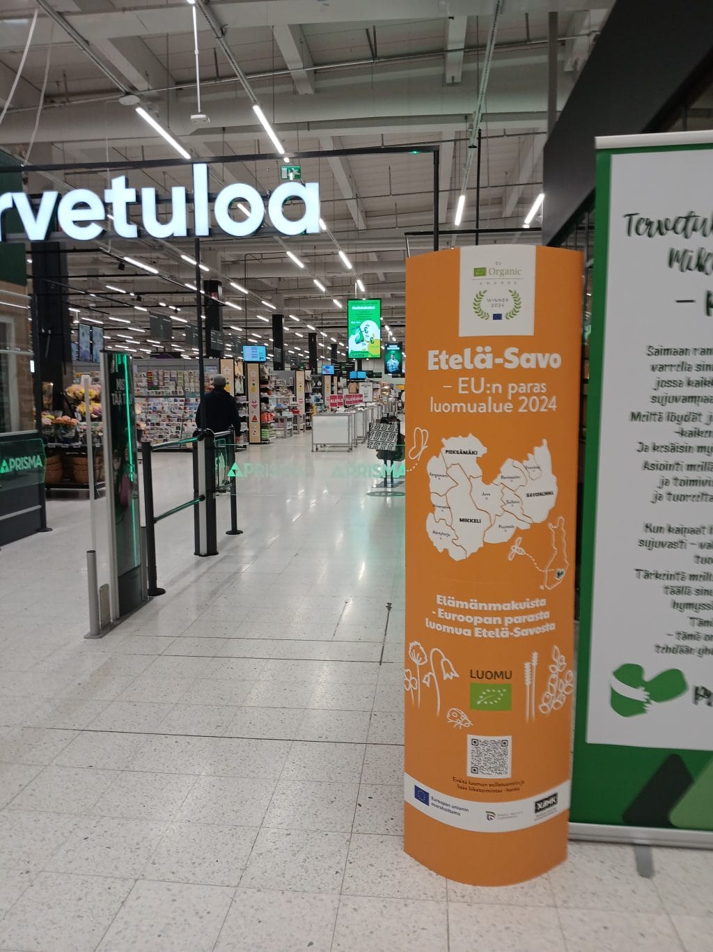 Kaupan sisätilan sisäänkäynti. Oikealla keltapohjainen korkea mainos, jossa Etelä-Savo piirroskuvana ja EU:n luomumerkki. Tekstinä Etelä-Savo EU:n paras luomualue 2024.