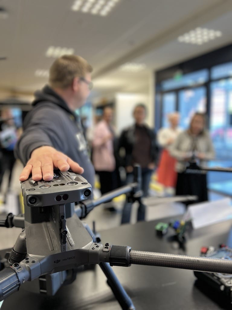 Mies pitää kättään dronen päällä, joka on pöydällä. Ihmisiä katsoo dronea dronen takaa.