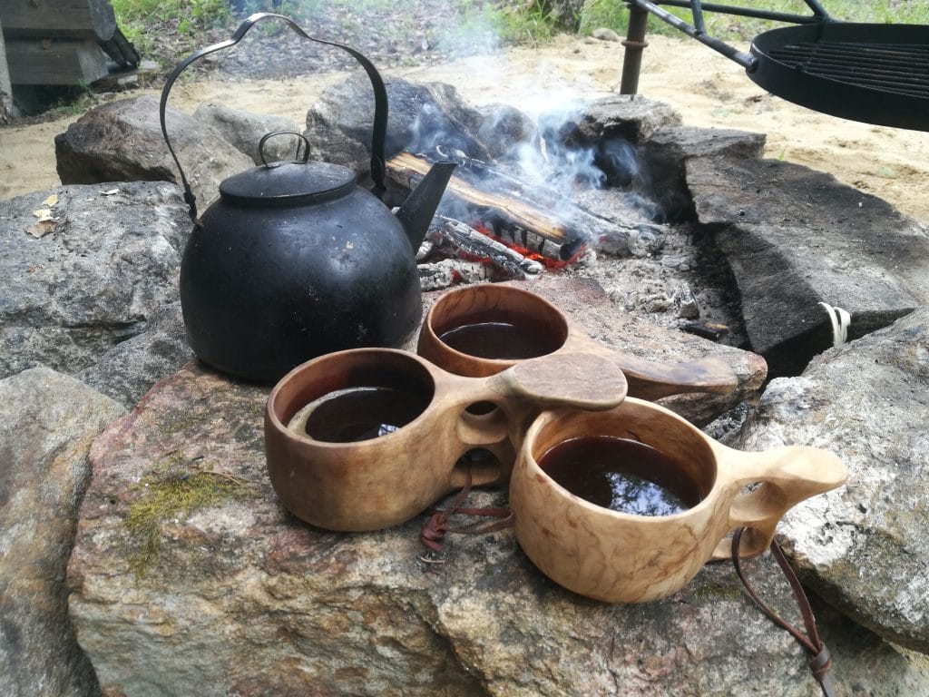 Kuvaassa on kivinen nuotiokehä, savuava nuotio. Kivikehän raunalla on musta kahvipannau ja kolem kuksaa, joihin on kaadettu kahvia