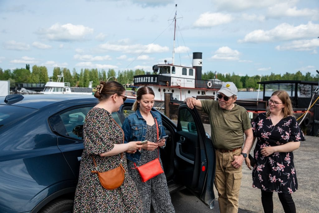Neljä ihmistä seisoo tumman auton vieressä. Kahdella vasemmanpuoleisella henkilöllä on kädessään puhelimet ja he katsovat niihin. Taustalla on järvi ja veneitä.