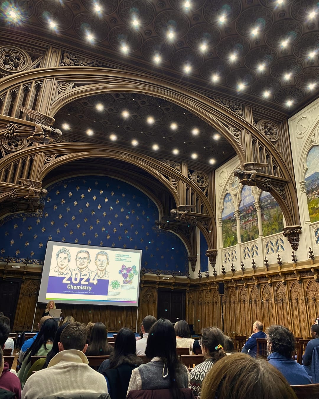 Opiskelijat istuvat vanhassa juhlasalissa, jossa on komeat puiset kaarikoristeet katossa.