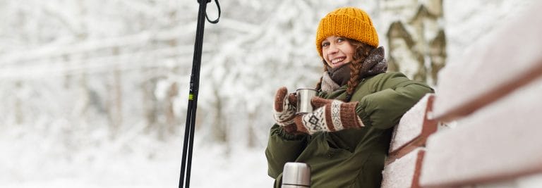 Nuori nainen istuu ulkona penkillä termosmuki kädessä lumisessa säässä.