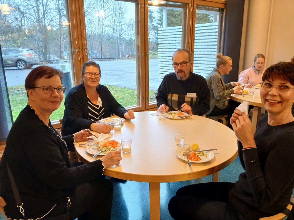 Kuvassa neljä ihmistä pyöreän pöydän äärellä syömässä koululounasta.