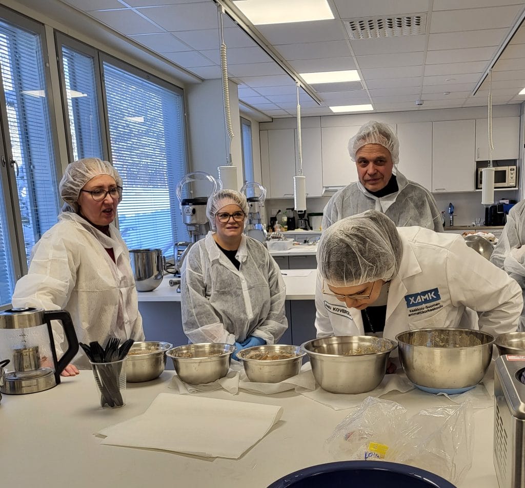 Ihmisiä suojatakeissa ja -myssyissä, yksi on kumartunut haistelemaan kulhossa olevaa ruokaa.