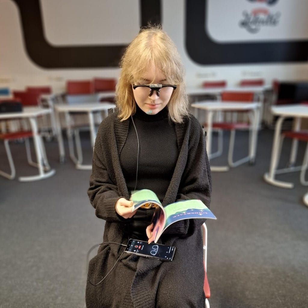 Kuvassa testihenkilö istuu tuolilla ja selailee lehteä. Päässään hänellä on puettava silmänliikekamera, joka seuraa hänen silmänliikkeitään.