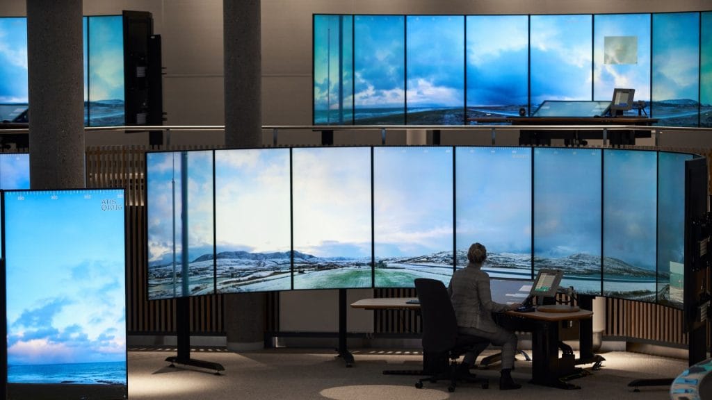 suuret digitaaliset näytöt, joihin heijastuu kuva lentokentän kiitoradasta. Näyttöjen äärellä istuu yksi henkilö.