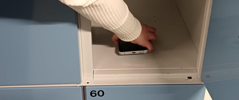 Valokuva. Ihmisen käsi asettamassa kännykkää säilytyslokeroon.