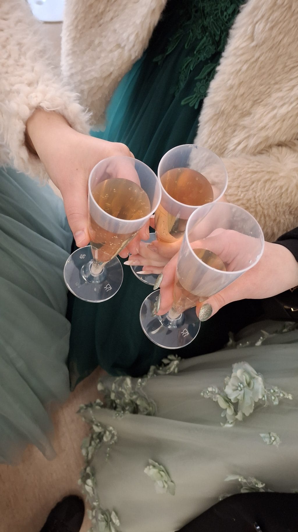 Kolme kättä kohottaa kuohuviinilaseja, joissa limsaa, lasien ympärillä näkyvät juhla-asut.