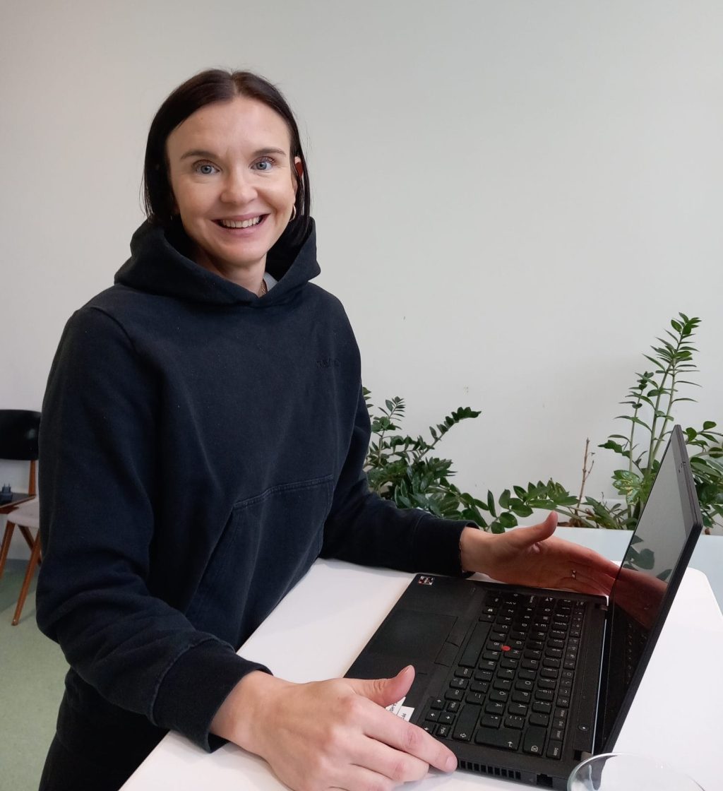 Henkilö istuu tietokoneen ääressä ja hymyilee kameralle