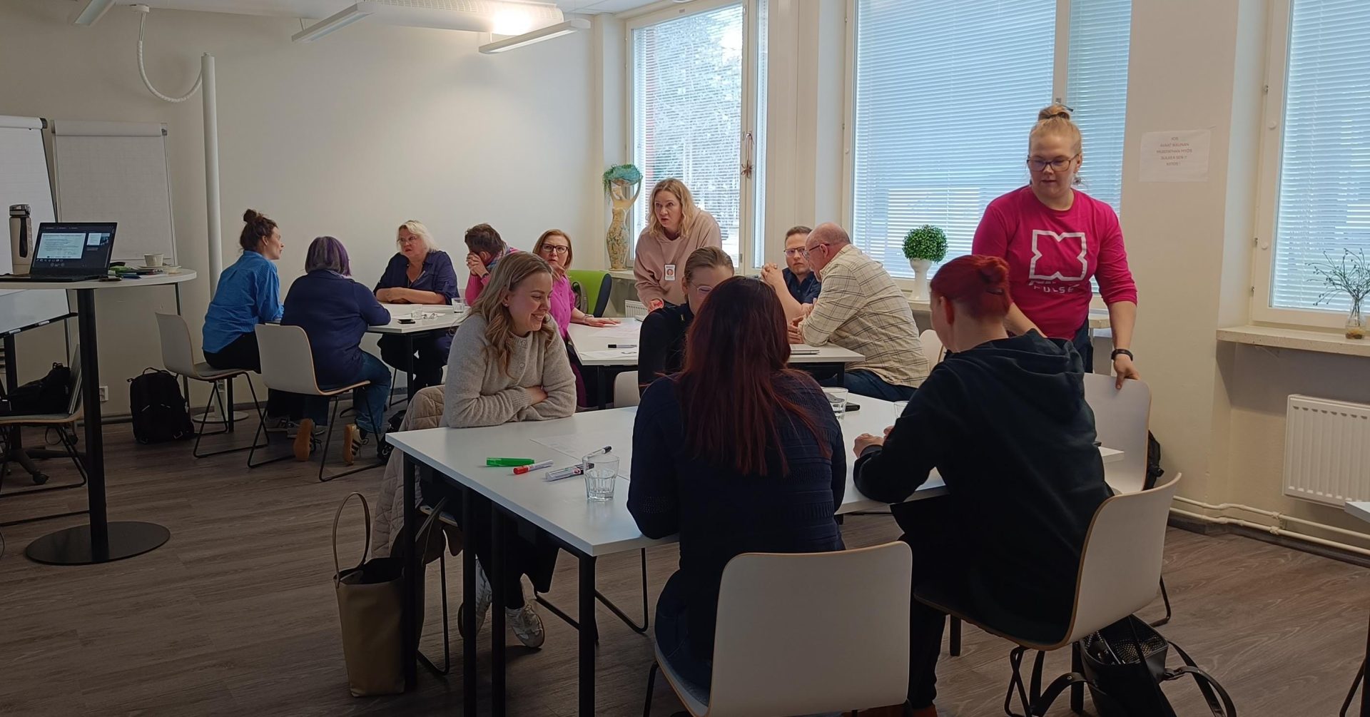 Luokkatilassa useita henkilöitä istuu pienryhmissä pöytien ääressä työskentelemässä. Etualalla neljä osallistujaa työskentelee yhdessä, pöydällä kyniä ja vesilasi. Taustalla muita ryhmiä, fläppitaulu ja esitystietokone. Ikkunoista tulee luonnonvaloa, ja yksi henkilö kävelee pöytien välissä.