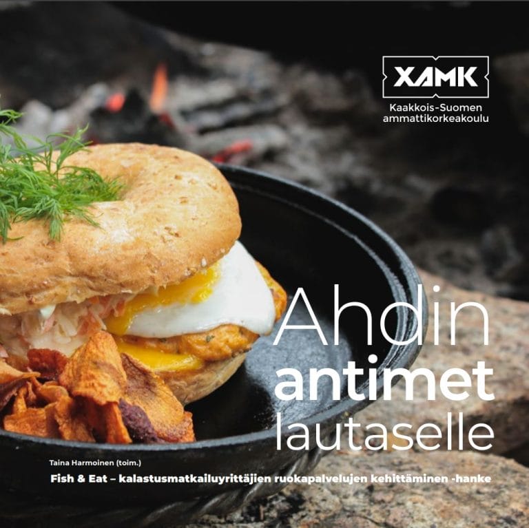 Mustalla pyöreällä pannulla sämpylä, jonka välissä täytteitä. Teksti Ahdin antimet lautaselle.