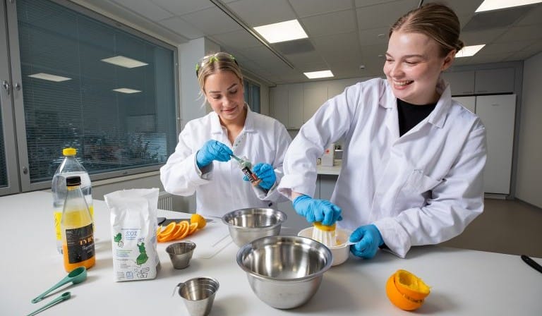 Kaksi valkotakkista nuorta naista työskentelee työpöydän ääressä. Pöydällä pyöreitä teräskulhoja ja ruoanlaittotarvikkeita.