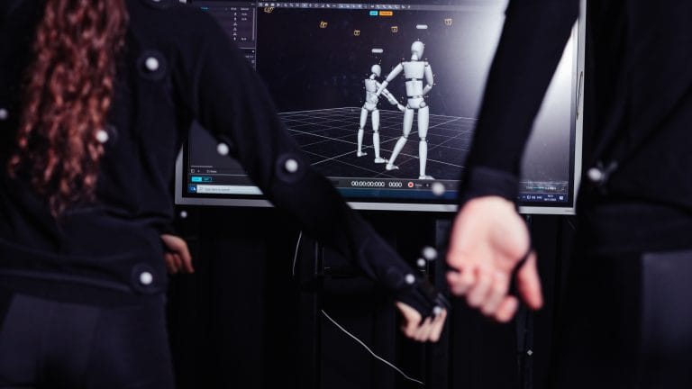 Motion capture session with two people in tracking suits and animated 3D figures displayed on a computer screen.