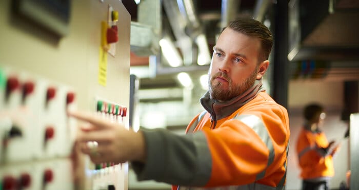 An industrial service engineer conducts a safety check of a control panel in a boiler room. He is wearing hi vis and is pressing one of the buttons on the control panel.