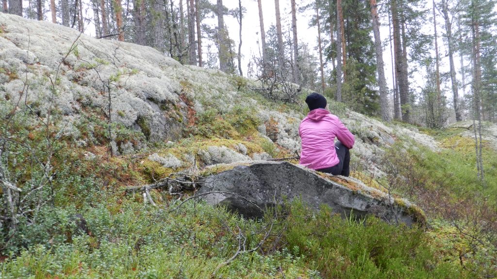 Henkilö istuu kivellä kallioisessa metsämaastossa vihreän kasvillisuuden ympäröivänä.