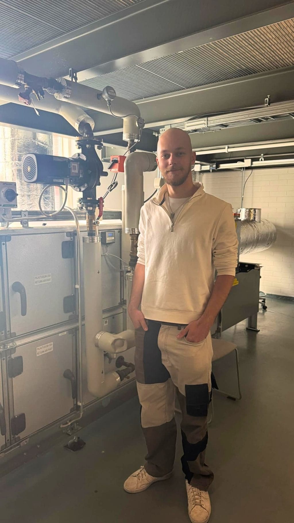 Henkilö seisoo talotekniikan laboratoriossa ilmanvaihtolaitteiston ja putkistojen vieressä.