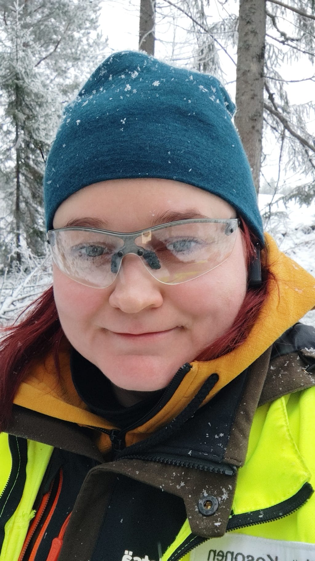 Henkilö seisoo lumisessa metsässä huomiovaatteissa, suojalasit ja sininen pipo päässään.