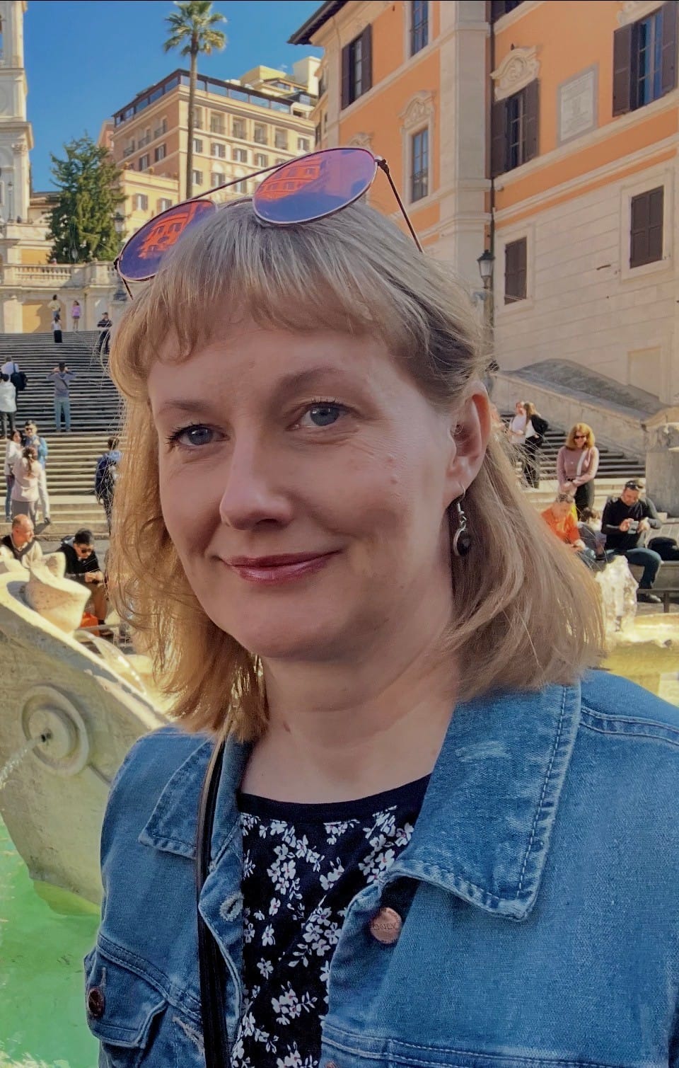 Lähikuva henkilöstä ulkona kaupunkiympäristössä. Taustalla näkyy vaaleita rakennuksia, portaat ja suihkulähde.