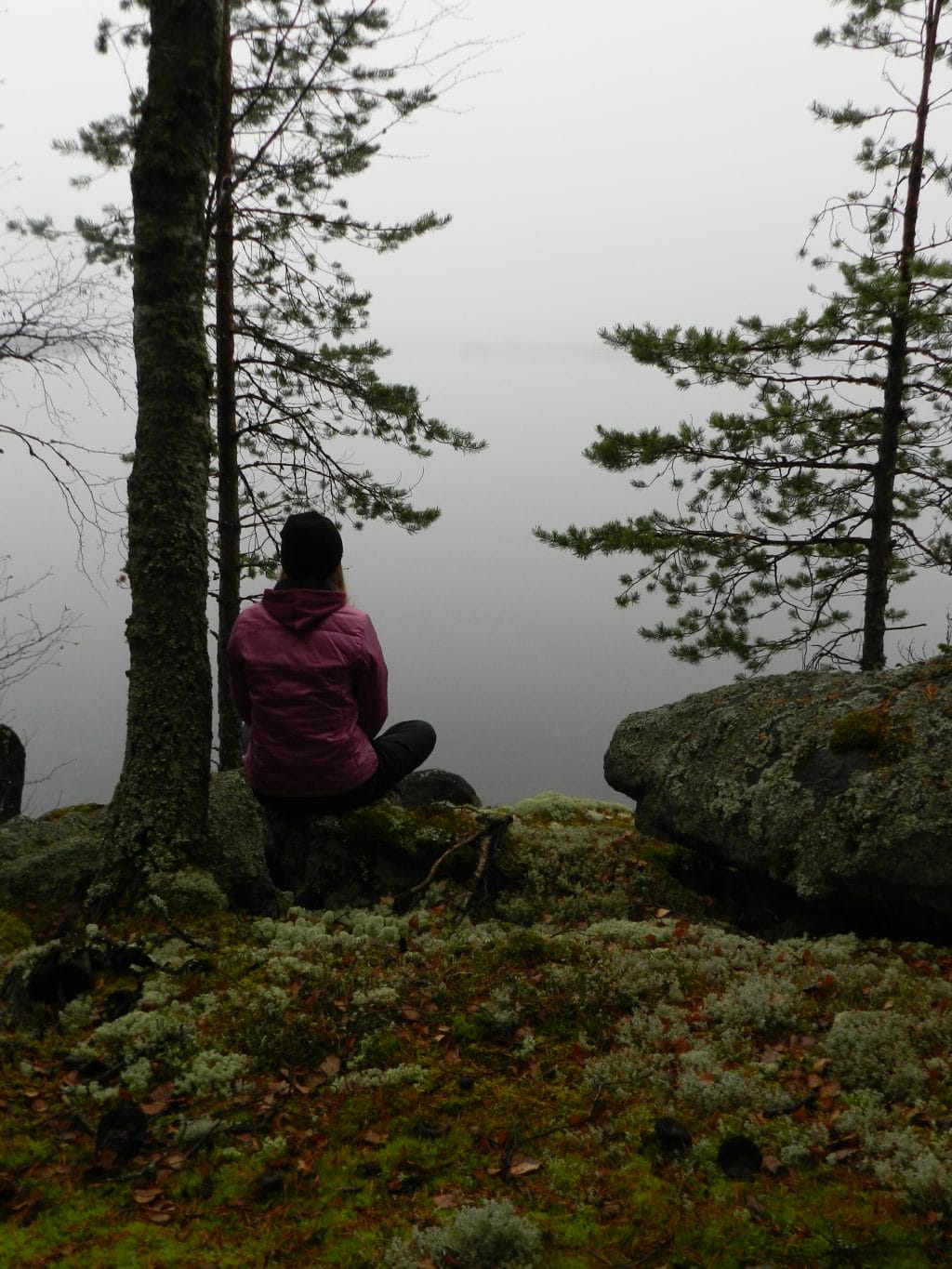 Henkilö istuu metsässä kallion reunalla veden äärellä havupuiden ja sammalpeitteisen maaston ympäröivänä.