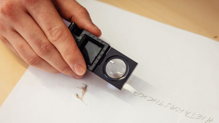 Käsi pitelee mittaluuppia paperille asetetun pienen kasvinäytteen päällä