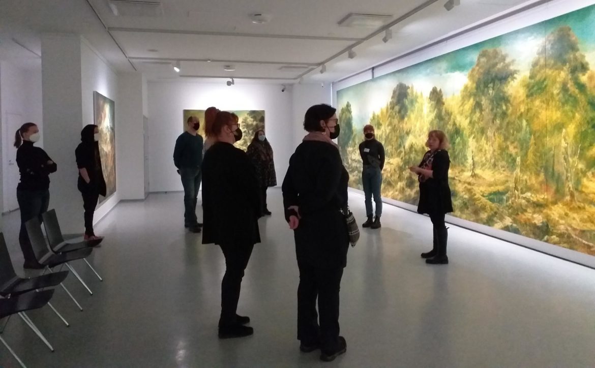 Ryhmä ihmisiä suuren maalauksen edessä kuuntelemassa opasten esittelyä taidenäyttelystä. 