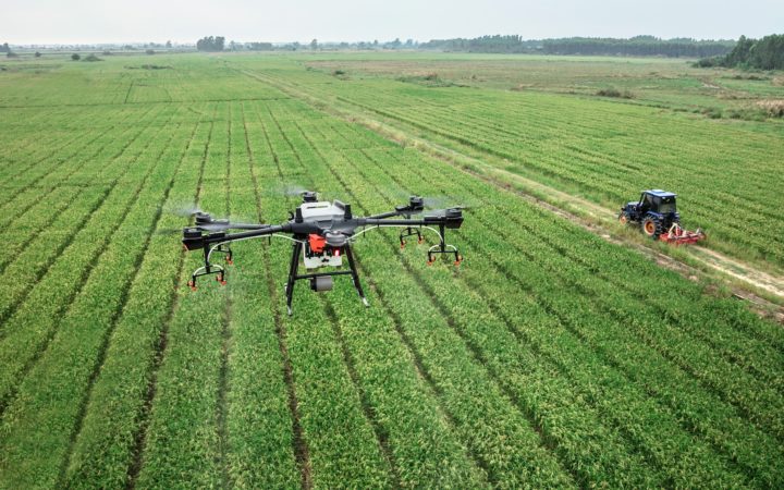 Kuvassa näkyy iso pelto, jonka päällä lentää drone. Alhaalla näkyy traktori.