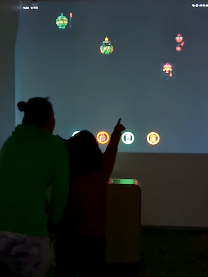 Kaksi henkilöä sytyttävät virtuaalisia lyhtyjä seinälle. Lapsi osoittaa seinälle projisoituja lyhtyjä.