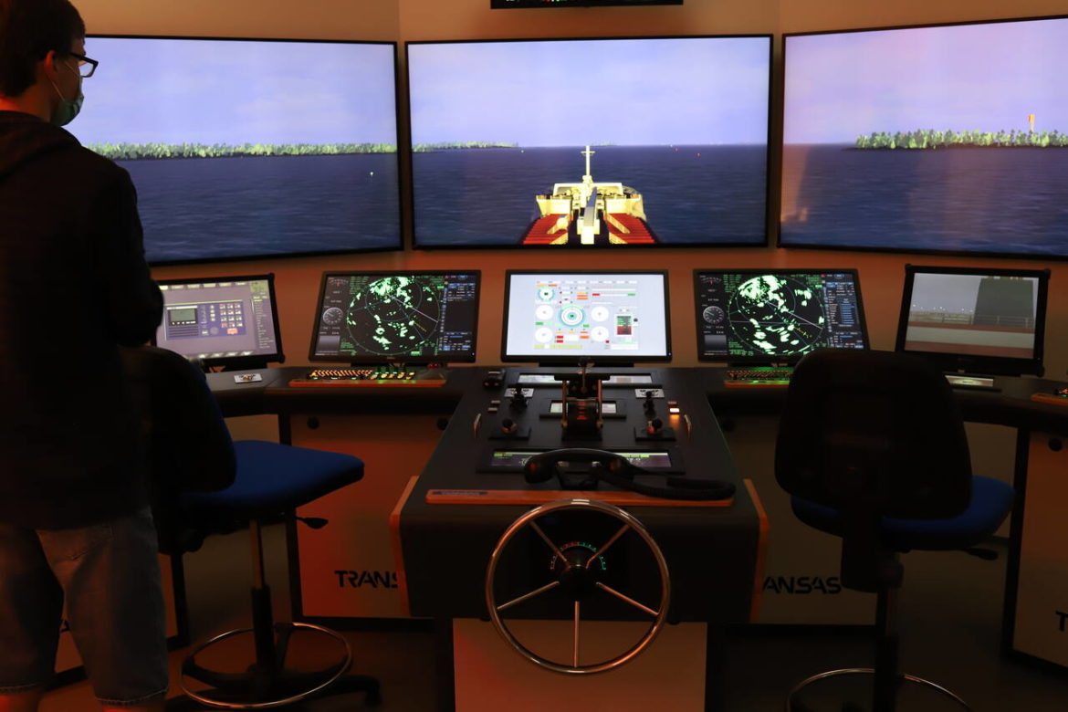 Mies kuvan vasemmassa reunassa merenkulun koulutuksen simulaattorinäyttöjen ja ohjaimien edessä.