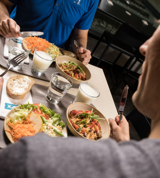 Ruokapöydässä kaksi vastakkain istuvaa henkilöä, joilla on tarjottimilla lautasilla ruoka-annokset.