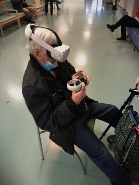 Iäkäs ihminen istuu käytävällä yllään virtuaalilasit ja käsissään ohjaimet